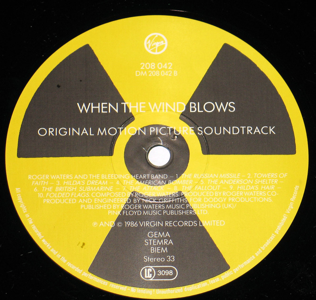 Record label of When the Wind Blows Original Motion Picture Soundtrack. The design features a bold yellow and black radiation hazard symbol in the background. At the top, the Virgin Records logo is displayed in white, along with the catalog number 208 042 and DM 208 042 B. The album title, When the Wind Blows, is printed in large white capital letters, followed by Original Motion Picture Soundtrack in a smaller font. Below, the tracklist for Side B is listed in white text, featuring compositions by Roger Waters and The Bleeding Heart Band, including The Russian Missile, Towers of Faith, Hilda’s Dream, The American Bomber, The Anderson Shelter, The British Submarine, The Attack, The Fallout, Hilda’s Hair, and Folded Flags. Credits mention that the music was composed and produced by Roger Waters and engineered by Nick Griffiths. Copyright details at the bottom indicate a 1986 release by Virgin Records Limited, with licensing and publishing information for Pink Floyd Music Publishers Ltd. and music rights organizations GEMA, STEMRA, BIEM. The overall design emphasizes the album’s anti-nuclear war themes with its stark contrast and warning symbol imagery